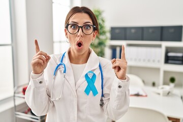 Wall Mural - Young brunette doctor woman wearing stethoscope at the clinic amazed and surprised looking up and pointing with fingers and raised arms.