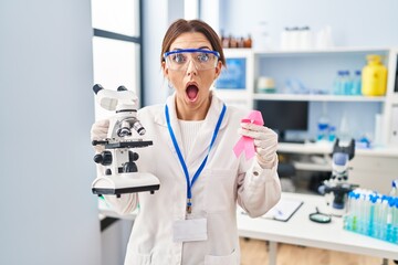 Sticker - Young brunette woman working at scientist laboratory holding pink ribbon afraid and shocked with surprise and amazed expression, fear and excited face.