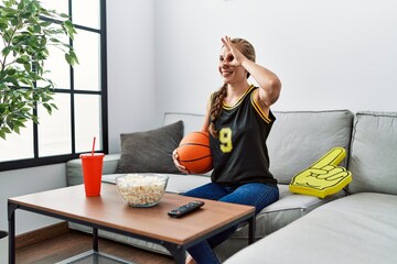 Poster - Young blonde woman holding basketball ball cheering tv game smiling happy doing ok sign with hand on eye looking through fingers