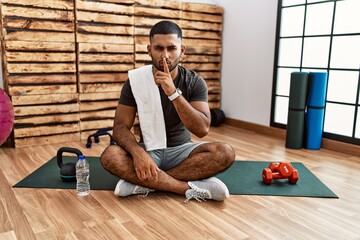 Canvas Print - Young indian man sitting on training mat at the gym asking to be quiet with finger on lips. silence and secret concept.