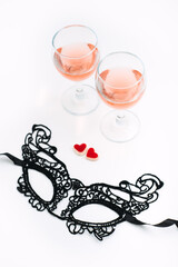 Valentines Day Festive table setting with black lace mask, two glasses of rose wine and two red heart shape candies on white table. Valentine Day, flirting, romance, dating concept, copy space