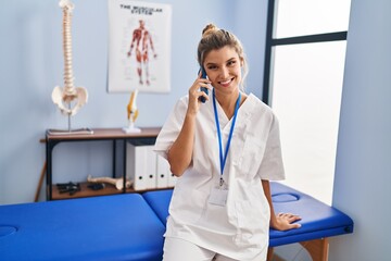 Poster - Young blonde woman wearing physiotherapist uniform talking on the smartphone at clinic