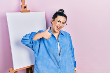 Poster - Young hispanic woman standing by painter easel stand doing happy thumbs up gesture with hand. approving expression looking at the camera showing success.