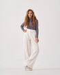 © Dmitry Tsvetkov - Pretty woman with stylish makeup and long flowing hair wearing sweater and loose trousers holds hand on hip on white background