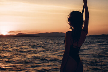 Wall Mural - Portrait of a lovely young skinny female model posing on her boat. Silhouette of a girl in a bikini on a sunset background. Against the backdrop of the raging sea and nature on the horizon. 