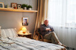 © pressmaster - Tired senior man sitting in armchair by window and napping in the middle of day in front of double bed in home environment