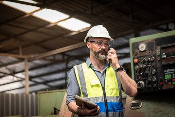 Caucasian male engineer or technician in safety uniform are standing working and checking maintenance machinery plan in tablet and talking by walkie talkie at industrial manufacturer factory.