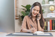 © KMPZZZ - Smiling, entrepreneur asian young professional business woman discussing on call mobile, using device laptop computer on desk, holding smartphone talking to client while sitting work at home office.