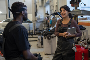 Wall Mural - Happy young Latin woman with tablet interacting with African male colleague during work in garage