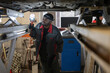 © pressmaster - Young African man in workwear and gloves holding lamp by lower part of car while standing under vehicle and checking it