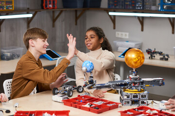 Wall Mural - Boy and girl congratulating each other with successful project, they sitting at the table with robotics objects