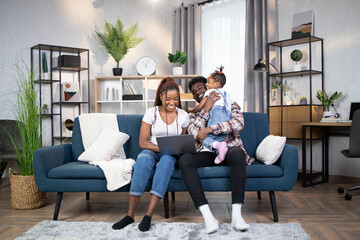 Wall Mural - Beautiful african woman typing on modern laptop while her husband and daughter playing together. Cheerful young family sitting together on comfy couch.