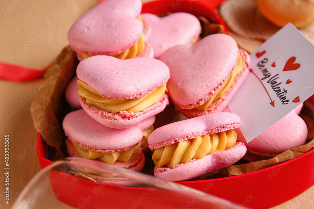 Box with tasty heart-shaped macaroons and greeting card on beige background