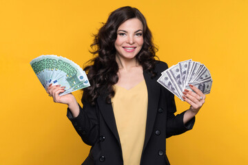 Young cheerful wealthy woman holding two cash fans of hundred euro and dollar banknotes of money, looking at camera on yellow background. Paper Currency Choice. Currency conversion. Investing concept.