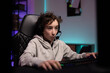 © ABCreative - Focused young boy with curly hair spends time at computer playing multiplayer video games with team, talks to people through headset, dark room night lit by neon led lights