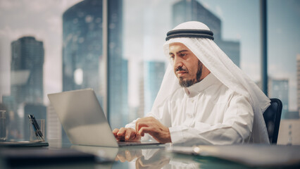 Wall Mural - Successful Arab Businessman in White Traditional Kandura Sitting in Office and Working on Laptop Computer. Business Manager Planning Investments. Saudi, Emirati, Arab Businessman Concept.