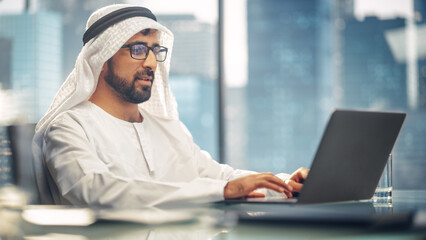 Wall Mural - Successful Arab Businessman in Glasses and White Traditional Outfit Sitting in Office and Working on Laptop. Business Manager Planning Corporate Strategy. Saudi, Emirati, Arab Businessman Concept.