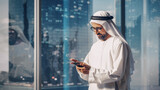Fototapeta  - Successful Muslim Businessman in Traditional White Outfit Standing in His Modern Office, Using Smartphone Next to Window with Skyscrapers. Successful Saudi, Emirati, Arab Businessman Concept.