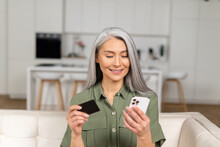 Serene Middle-aged Grey-haired Asian Woman Shopping Online Sitting With Credit Card And Mobile Phone At Home, Multiracial Senior 40s Female Making Order Online, Booking, Transfering Money