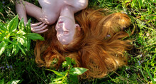 Beautiful Young Sexy Redhead Woman Portrait, Red Hair Spread In The Flowers, Lying Relaxed On The Green Meadow With Bare Shoulders, Enjoying Summer Sun And Fresh Air