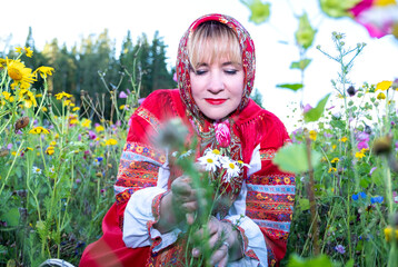 A young blonde Caucasian woman in a red national dress and with a red shawl. A woman is standing in a field of flowers, smiling and looking at the camera.