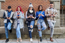 Full-body Multiethnic Men And Women In Casual Clothing Using Cell Phones While Leaning Against A Brick Wall On A City Street