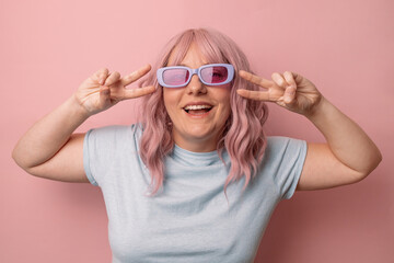 Smiling woman posing in glasses makes v sign shows peace gesture isolated over pink background.