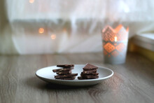 Plate With Almond Chocolate Pieces On The Table. Lit Candle And Bokeh Lights In The Background. Selective Focus.