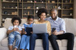 © fizkes - African spouses and children sit on sofa enjoy leisure use diverse electronic gadgets. Kids absorbed in videogame, instead live communication prefer online fun, bad life habit, devices overuse concept