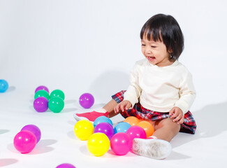 Wall Mural - Studio shot of little cute short black hair Asian baby girl daughter model in casual plaid skirt sitting on floor smiling laughing playing with colorful round balls toy alone on white background