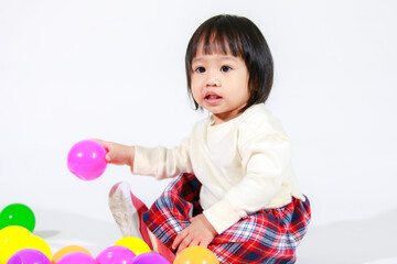 Wall Mural - Studio shot of little cute short black hair Asian baby girl daughter model in casual plaid skirt sitting on floor smiling laughing playing with colorful round balls toy alone on white background