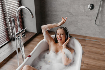 Wall Mural - Happy relaxed lady taking bath full of foam and listening to music in wireless headphones, singing and dancing