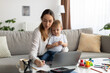 © Prostock-studio - Motherhood and education. Young lady with baby on hands studying online on laptop and taking notes to workbook