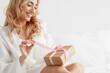 © Prostock-studio - Glad young attractive european blonde female in robe, opens gift box, unties ribbon on white bed