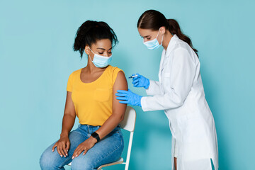Poster - Female doctor making injection of coronavirus vaccine to young black lady in protective mask, blue studio background