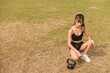 A slim and petite Filipino woman rests and kneels while looking at her kettlebell. At an grassy open field. Outdoor exercise lifestyle.