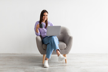 Wall Mural - Positive Caucasian woman working online, sitting in armchair with coffee and using laptop against white studio wall