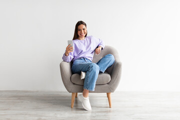 Poster - Smiling millennial woman chatting online on smartphone, sitting in comfy armchair on white studio background