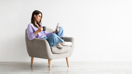 Poster - Young Caucasian woman with coffee sitting in armchair and using tablet computer against white studio wall