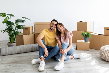 Wall Mural - Relocation concept. Happy multiracial homeowners sitting on floor of their property among carton boxes, full length