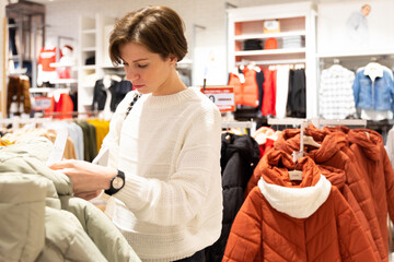 Wall Mural - Photo of young attractive brunette woman with a short haircut in a white sweater chooses stylish and casual clothes in a store in a shopping centre