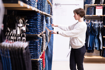 Wall Mural - Photo of young attractive brunette woman with a short haircut in a white sweater chooses stylish and casual jeans and denim clothes in a store in a shopping centre