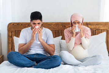 Wall Mural - Muslim pregnant couple got flu, having runny nose, sitting on bed and blowing nose, sneezing in paper tissue