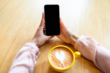 Poster - Closeup of millennial lady holding smartphone with blank black screen at table in cafe, drinking coffee, mockup