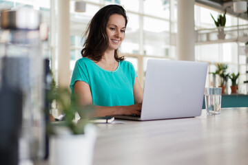 happy smiling remote working dark haired woman typing on laptop or notebook in casual outfit sitting on her work desk in her modern bright loft living room home office with many windows