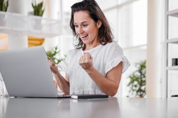 happy joyful successful remote working woman cheerful with fists infront of laptop or notebook in casual outfit sitting on work desk in modern loft living room home office