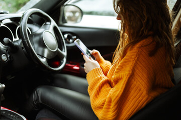 Portrait of a young casual woman in orange sweater, texting on her smartphone or making video call ,while driving or traffic stop a car during lunch break.