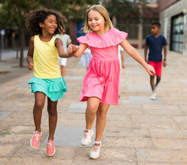 Happy blonde preteen girl enjoying walk with african american girl friend along city street on summer day, running skipping gaily and holding hands. Childhood friendship concept