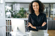 © Kateryna - Portrait of successful influential pretty African American businesswoman, company leader, manager, standing in her modern office near the workplace, with arms crossed, looks at camera, smiles friendly