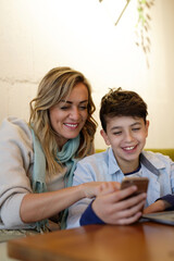 Mother and her young teenage son browsing internet content or social media on the phone, having fun together in a coffee shop